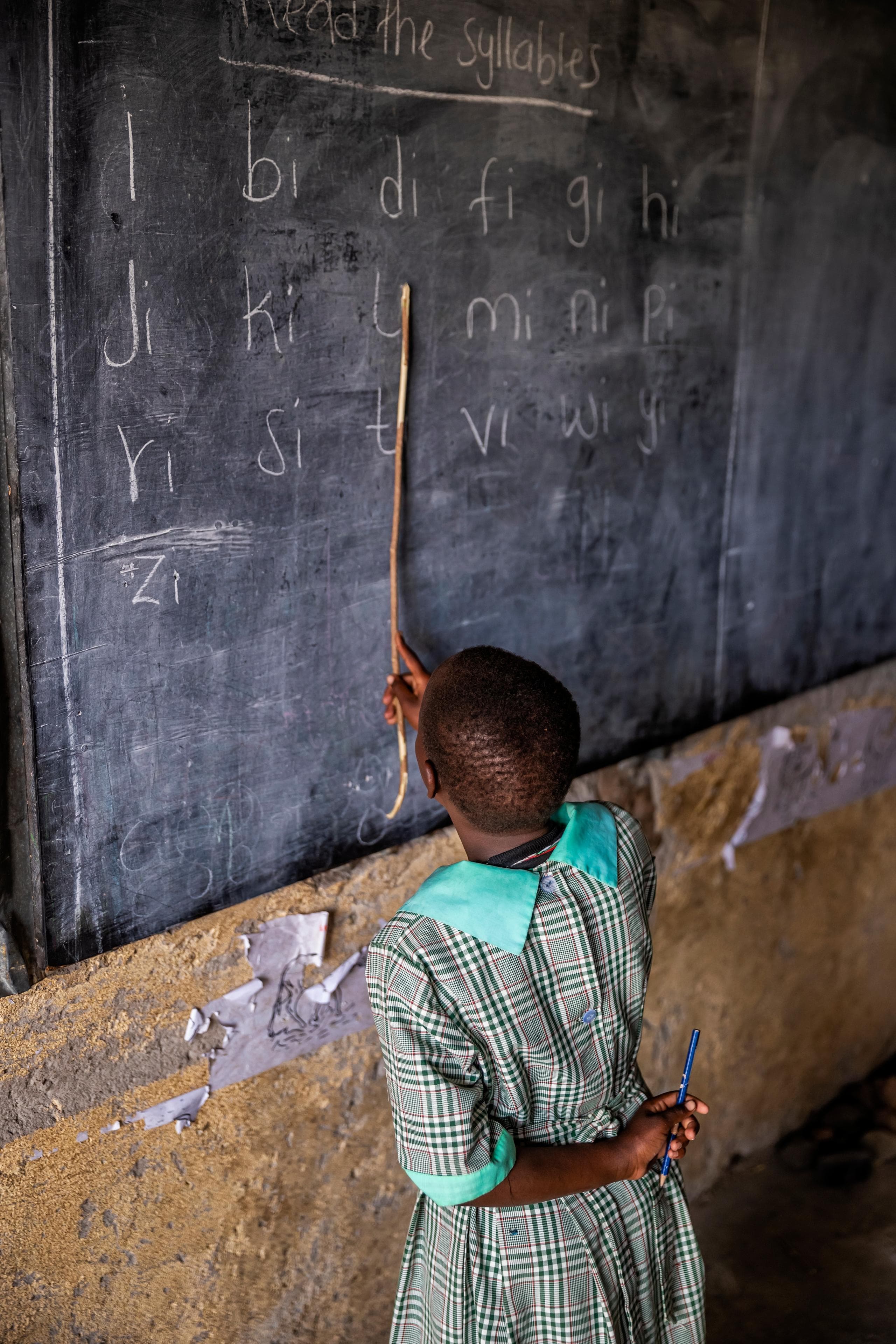 A girl in a school uniform standing in front of a chalk board pointing at text with a stick.