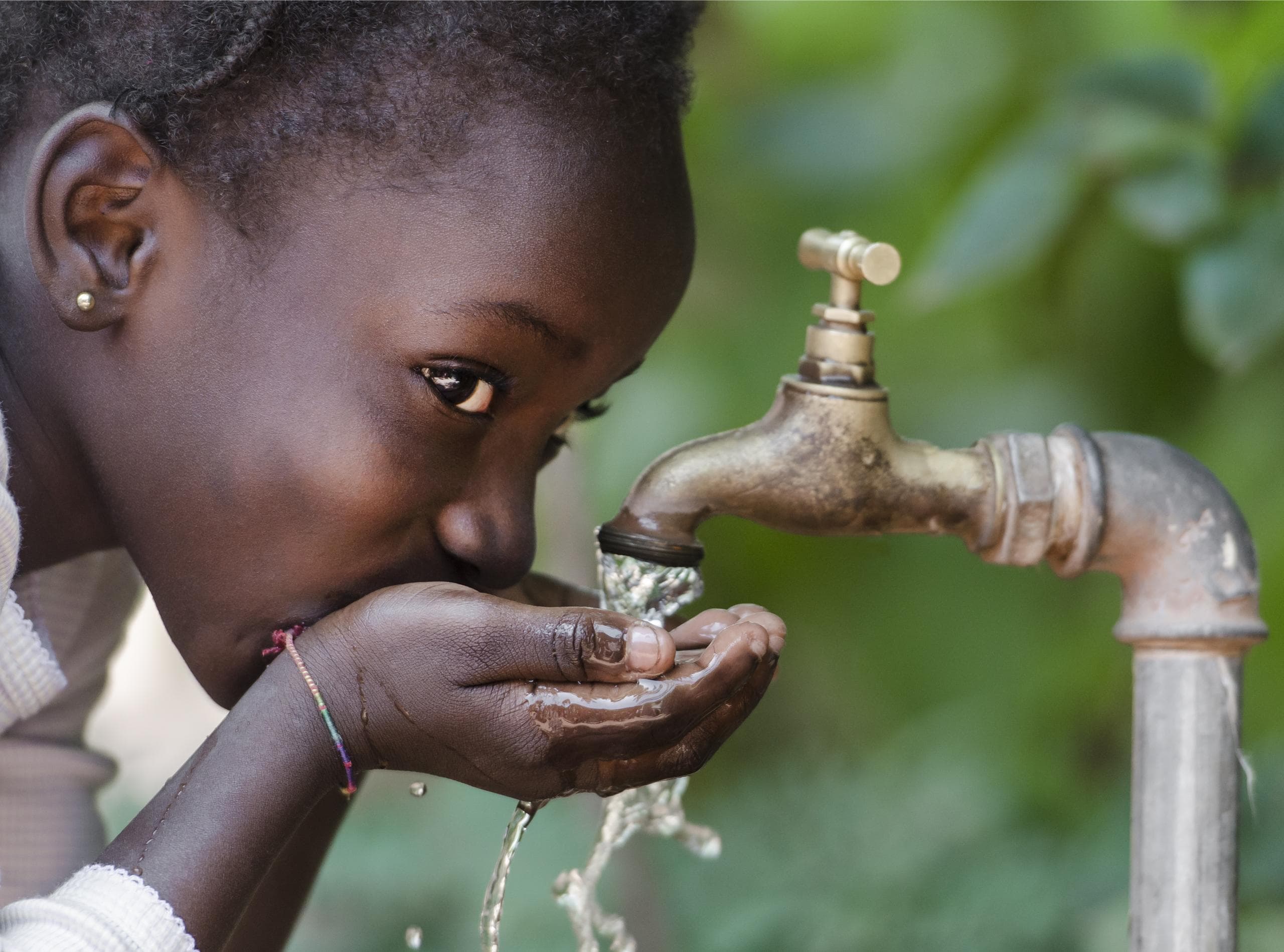 A girl dressed in white drinking water from an outdoor faucet.